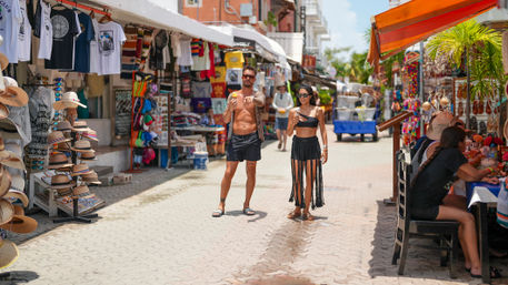 Tourists in swimwear enjoy ice cream while strolling a sunny coastal pedestrian market street lined with colorful souvenir stalls, hats and t-shirts
