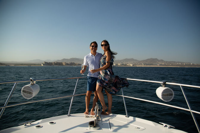 Couple standing on the bow of a yacht holding drinks, sunlit deep-blue ocean with a distant coastal city and low mountains on the horizon — relaxed seaside vacation.