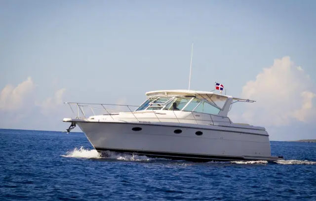 White motor yacht cruising across deep blue ocean, slicing small waves under a sunny sky with a small red-white-blue flag flying from the stern.