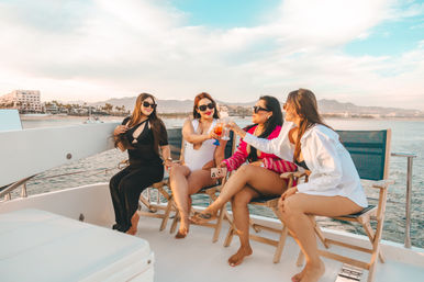 Four women in swimsuits and sunglasses toasting cocktails on a yacht deck with ocean, marina buildings and distant mountains under a golden-hour coastal sky