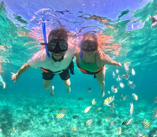 Two snorkelers in clear turquoise tropical water above a shallow coral reef, smiling amid a school of striped tropical fish