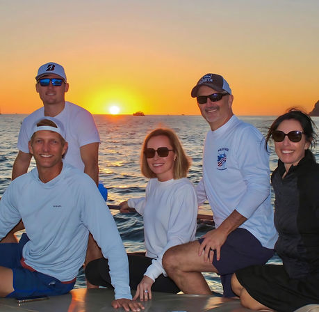 Five adults in sunglasses enjoying a boat ride at an ocean sunset, golden sun dipping below the horizon with calm water and a distant yacht on the water.