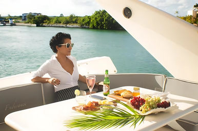 Woman in sunglasses lounging on a luxury yacht deck overlooking a turquoise tropical harbor, enjoying a brunch spread of charcuterie, bread, grapes and drinks.