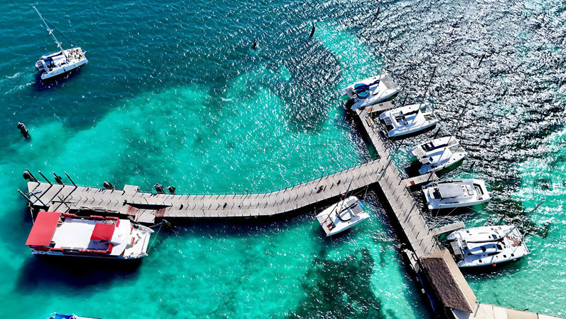 Aerial view of a curved wooden pier with docked yachts and a red-canopy tour boat on sparkling turquoise tropical water.