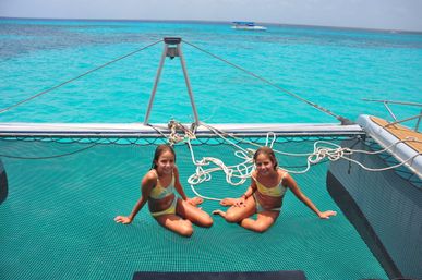 Two smiling girls in matching yellow swimsuits sitting on a catamaran trampoline over vivid turquoise tropical sea with coiled ropes and a boat on the horizon.