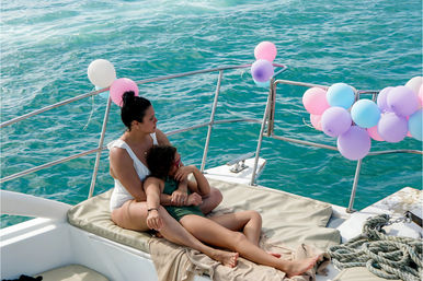 Sunlit woman and child cuddling on a yacht sunpad surrounded by pastel pink, purple and blue balloons with turquoise ocean waters in the background.