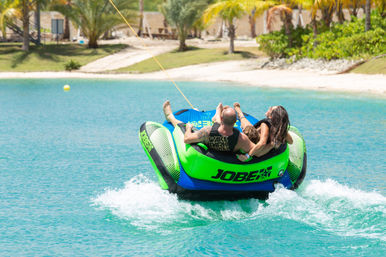 Family tubing on a bright green inflatable in clear turquoise water off a palm‑fringed tropical beach, being towed behind a boat