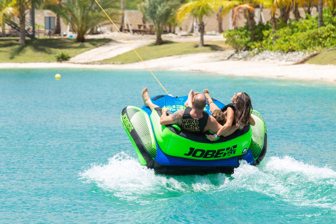 Family tubing on a bright green inflatable in clear turquoise water off a palm‑fringed tropical beach, being towed behind a boat
