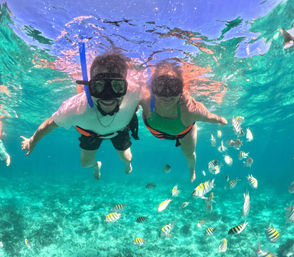 Two snorkelers in clear turquoise tropical water above a coral reef, smiling through masks and surrounded by striped yellow-and-black tropical fish.