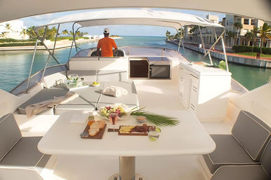 Luxury yacht deck with table set for brunch—charcuterie, drinks and cushions—person in orange at the helm cruising a palm-lined turquoise waterfront canal past seaside condos.