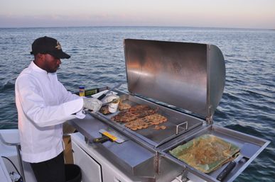 Person grilling skewered shrimp and lobster tails on a stainless-steel griddle aboard a boat, calm open ocean and pastel sky in the background — a sizzling seafood feast at sea.