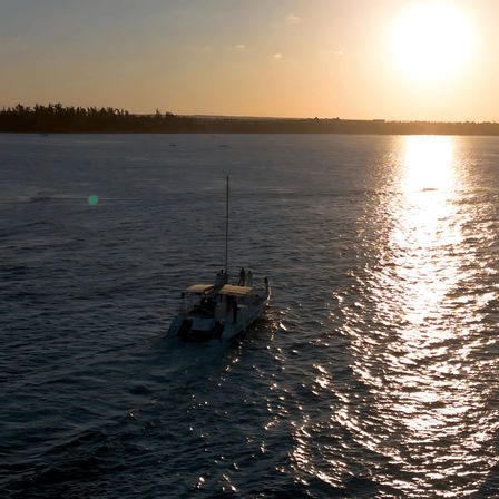 Small boat cruising across rippled ocean toward a tree-lined coastline at golden sunset, sun casting a bright shimmering reflection on the water.