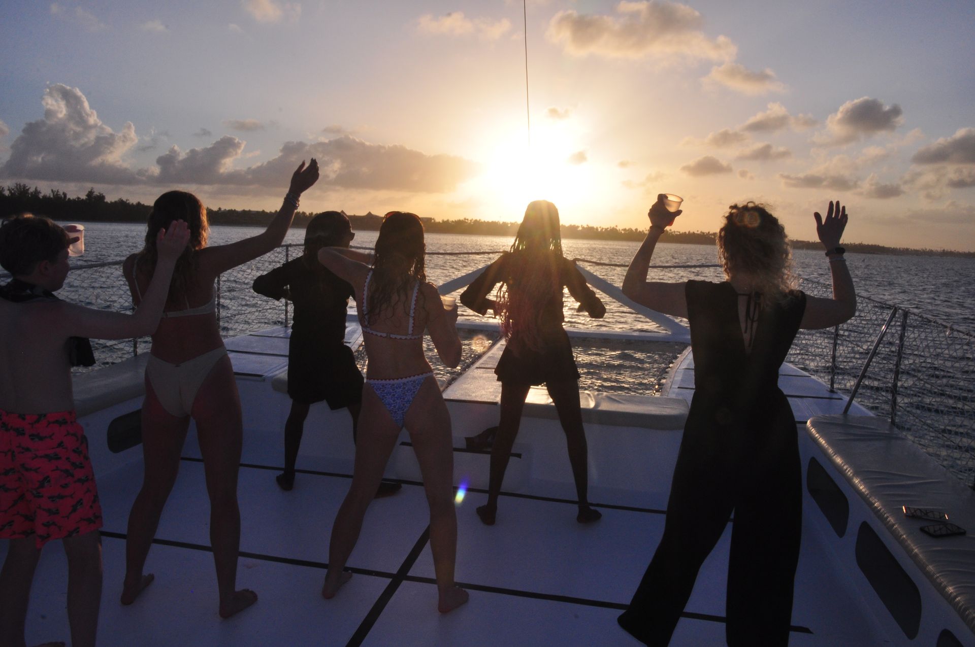 Silhouetted group of friends dancing and raising cups on a catamaran deck at a golden tropical sunset with island coastline and calm ocean