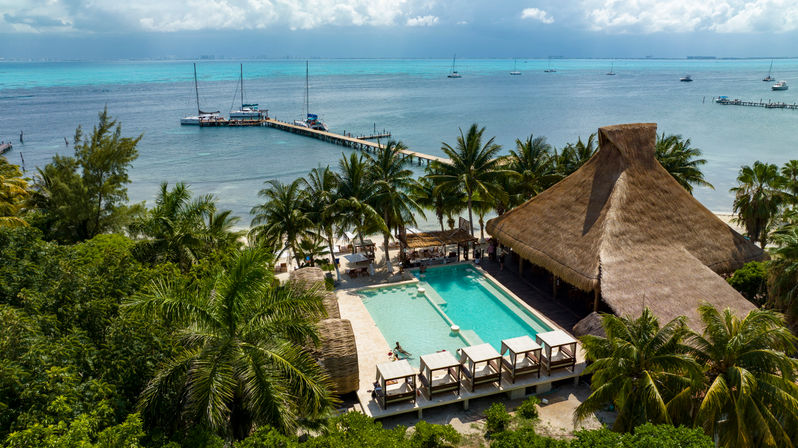 Aerial view of a sun-soaked tropical beach resort with turquoise ocean and sailboats, long wooden pier, palm-fringed white sand, a lagoon-style pool with cabanas and a large thatched-roof palapa