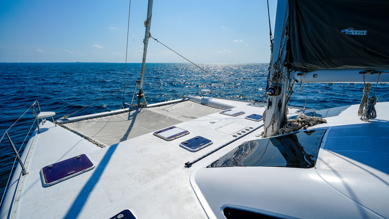 Catamaran bow with trampoline net, rigging and coiled ropes cruising on sparkling blue ocean under a clear sunny sky and distant horizon.