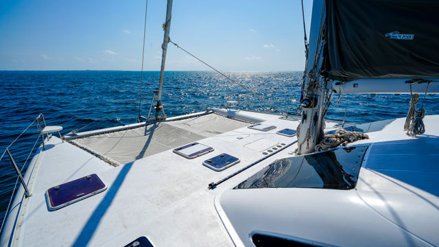 Catamaran bow with trampoline net, rigging and coiled ropes cruising on sparkling blue ocean under a clear sunny sky and distant horizon.