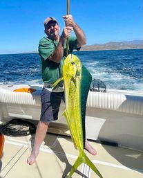 Angler on a boat holding a giant bright yellow-green mahi-mahi (dolphinfish) over the deck with clear blue offshore ocean and distant coastline — trophy sport-fishing catch.