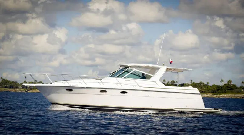 Sleek white motor yacht cruising on deep blue ocean past a palm-lined tropical shoreline under a bright sky with puffy clouds.