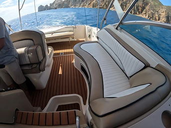 Onboard view of a motorboat with curved white-and-tan leather seating and teak deck, cruising on deep blue ocean toward rugged coastal cliffs under a sunny sky.
