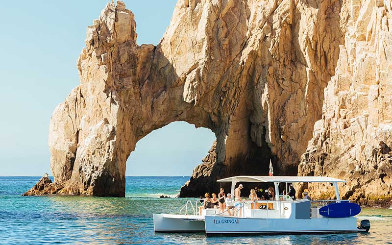 Tour boat with passengers cruising turquoise waters beside a dramatic natural rock arch at Cabo San Lucas on a sunny day.