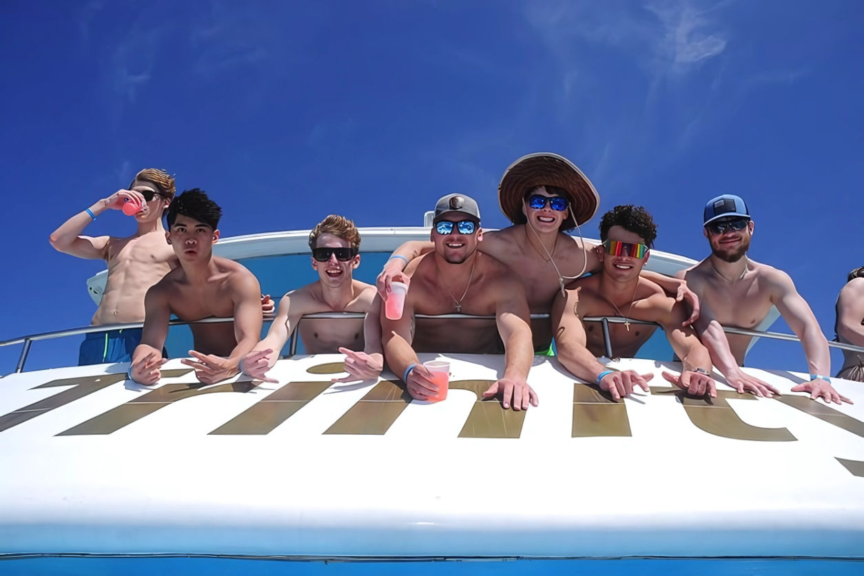 Group of shirtless young men leaning over a boat railing, smiling and holding colorful drinks under a bright blue sky during a sunny summer yacht party