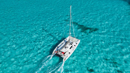 Aerial view of a white catamaran sailing through clear turquoise tropical water, twin hulls cutting foamy wakes with passengers relaxing on the sunlit deck.