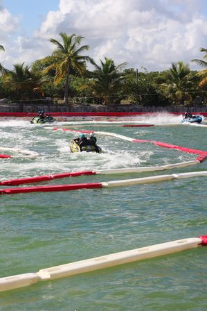 Three jet skis with helmeted riders racing inside red-and-white safety buoys on turquoise lagoon water, palm trees and a partly cloudy tropical sky in the background