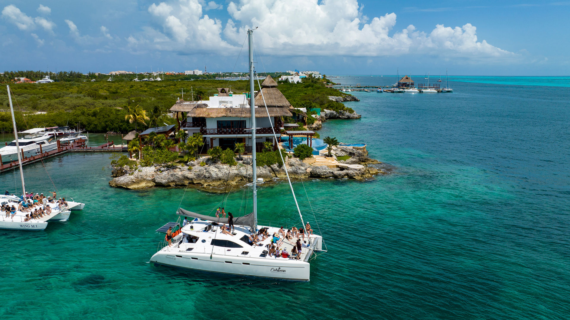 Catamaran tour with passengers anchored in turquoise Caribbean waters beside a rocky islet topped with a thatched-roof seaside villa and palm trees under a partly cloudy sky