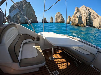 Sunlit boat deck with cushioned seating and table looking toward dramatic limestone sea stacks and a natural rock arch over turquoise ocean under a clear blue sky.