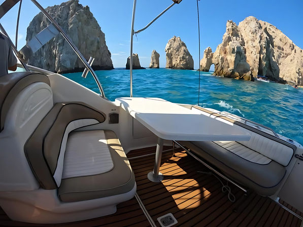 Sunlit boat deck with cushioned seating and table looking toward dramatic limestone sea stacks and a natural rock arch over turquoise ocean under a clear blue sky.