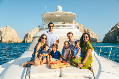 Four adults and four children smiling on the bow of a luxury yacht, enjoying a charcuterie board and champagne under sunny skies with rocky coastal cliffs in the background.