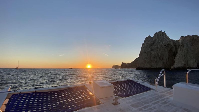 View from a catamaran deck with blue net trampolines in the foreground, sailboats on the sea and rugged coastal cliffs to the right as the sun sets over the ocean.