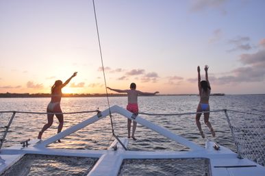 Three people leaping from the bow of a catamaran into calm ocean waters at sunset, arms raised against a pastel sky with a tropical coastline on the horizon — playful sunset cruise scene.