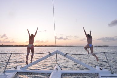 Two people jumping off the bow of a catamaran into the ocean at sunset with a tropical coastline silhouette, calm water and vacation vibes.