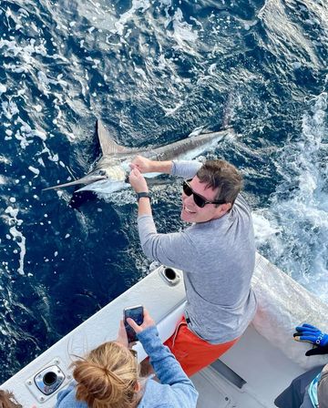 Smiling angler on a sportfishing boat holding a hooked billfish (marlin/sailfish) alongside the rail while a friend snaps a photo, choppy deep-blue offshore water in the background.