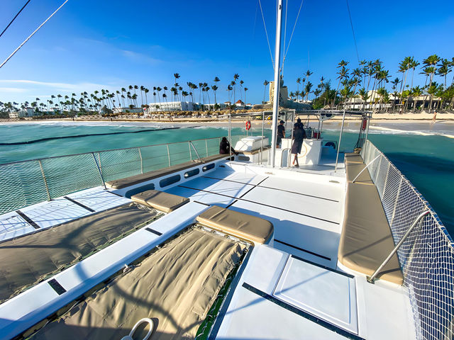 Sunlit catamaran deck with cushioned loungers and mesh nets overlooking turquoise water and a palm‑lined tropical beach under a clear blue sky