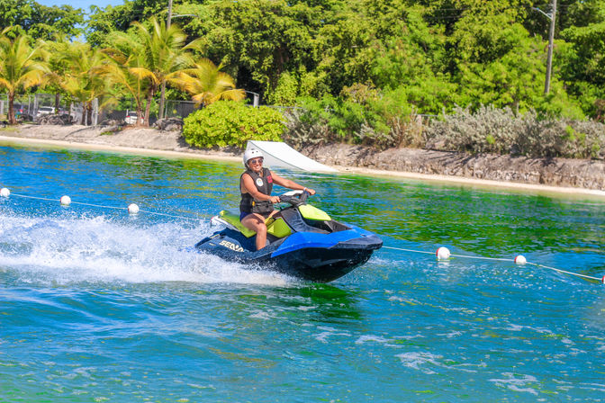 Person in helmet and life vest riding a blue and green jet ski across turquoise coastal water, spraying white wake past a palm-lined tropical beach on a sunny day.