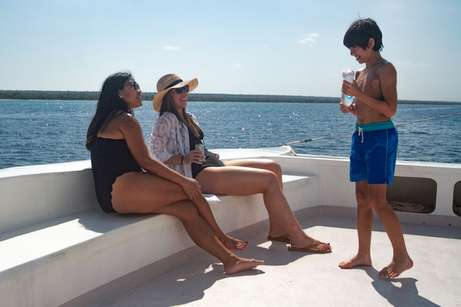 Two women relaxing on a white boat deck on a sunny day—one in a straw hat and cover-up—while a smiling boy in blue swim trunks holds a water bottle, with sparkling ocean and distant shoreline beyond.