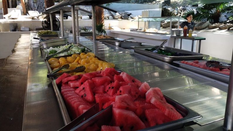 Outdoor buffet salad bar with stainless-steel trays of bright sliced watermelon, mandarin orange segments, lettuce and salad toppings under a glass sneeze guard, tongs in place in a tropical poolside setting.