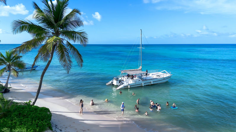 Tropical island beach with white sand, leaning palm trees and turquoise water; a white catamaran anchored near shore while people swim and relax under a bright blue sky.