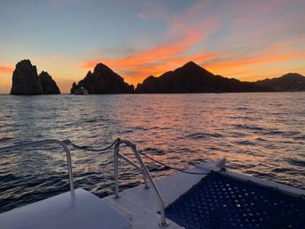 Boat deck view at sunset of rugged coastal rock formations with an orange-pink sky reflecting on calm ocean water