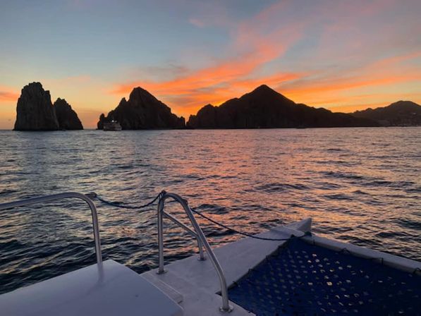 Boat deck view at sunset of rugged coastal rock formations with an orange-pink sky reflecting on calm ocean water