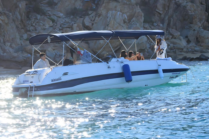 White motorboat with navy canopy carrying passengers taking selfies near a rocky Cabo coastline, sparkling turquoise water and sunlit waves