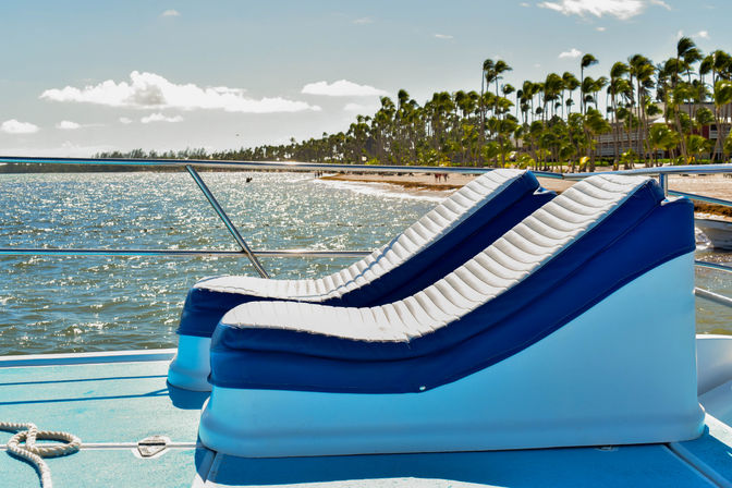 Two blue-and-white cushioned lounge chairs on a sunlit boat deck overlooking sparkling tropical ocean and a palm-lined sandy beach under a clear sky