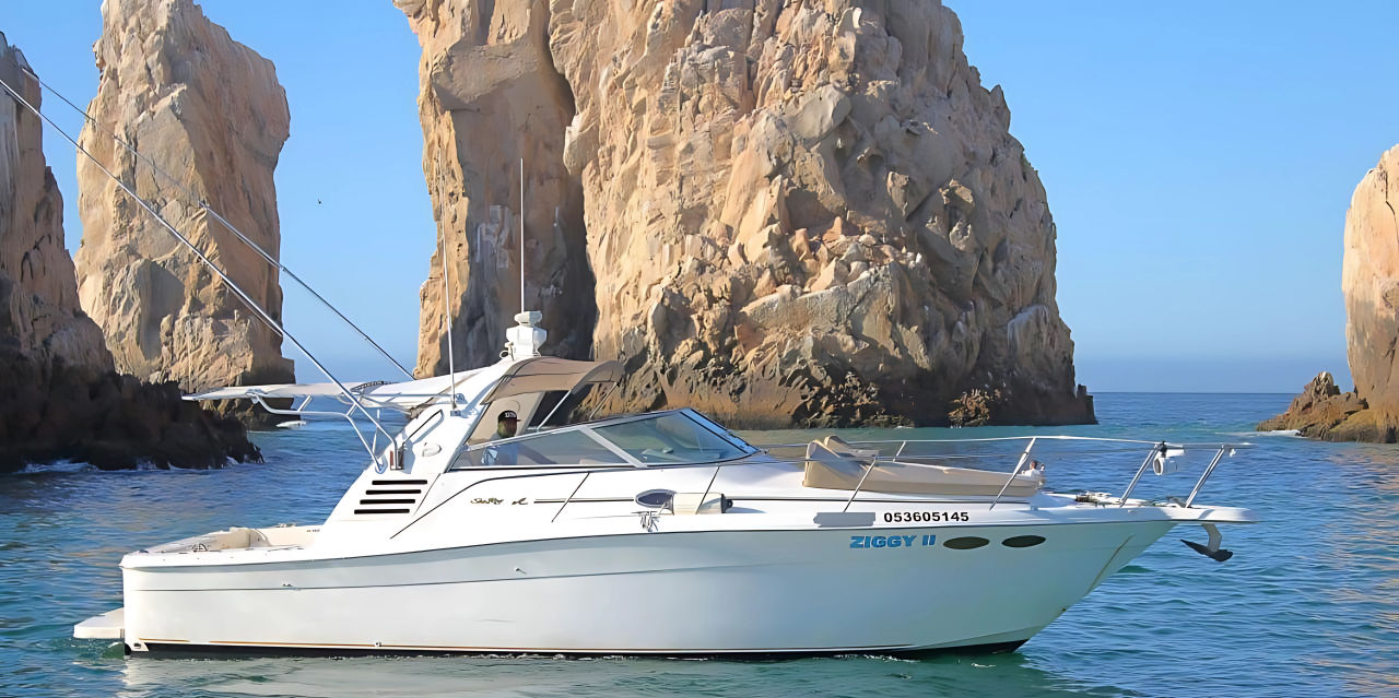 White motor yacht cruising turquoise water near towering sunlit granite sea stacks and a natural sea arch under a clear blue sky.