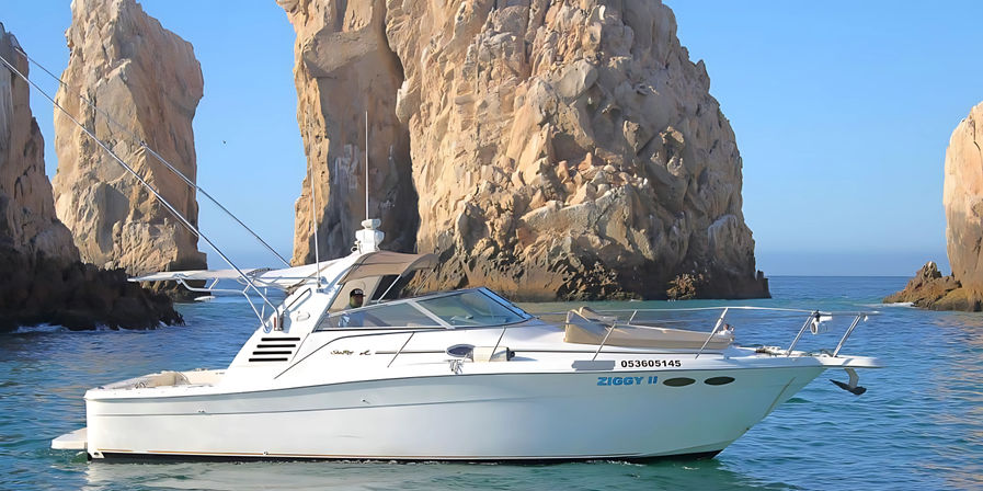 White motor yacht cruising turquoise water near towering sunlit granite sea stacks and a natural sea arch under a clear blue sky.