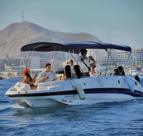 Group of people enjoying a sunny motorboat cruise under a navy canopy near a coastal resort harbor with waterfront hotels and hills in the background.