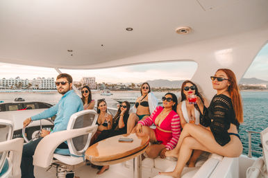Group of friends on a luxury yacht near a beachfront resort marina at sunset, wearing swimwear and sunglasses and sipping cocktails with coastal hotels and mountains in the background.