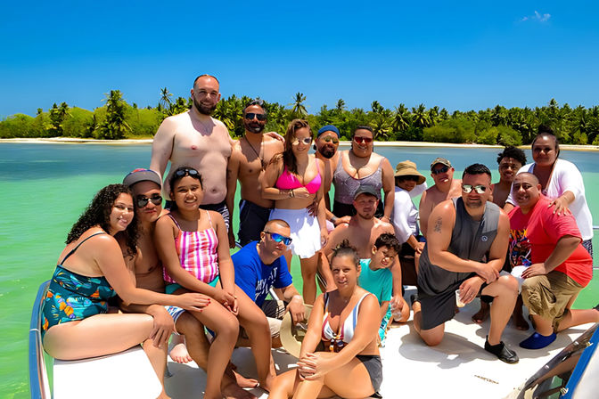 Large group of friends and family posing on a boat in a turquoise tropical lagoon with palm-fringed island shoreline and a bright blue sky — sunny vacation vibe.