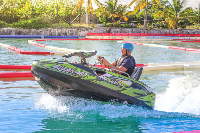 Rider in a blue helmet on a green personal watercraft cutting through turquoise lagoon water, splashing past red-and-white floating course barriers with palm trees on a sunny tropical shoreline.